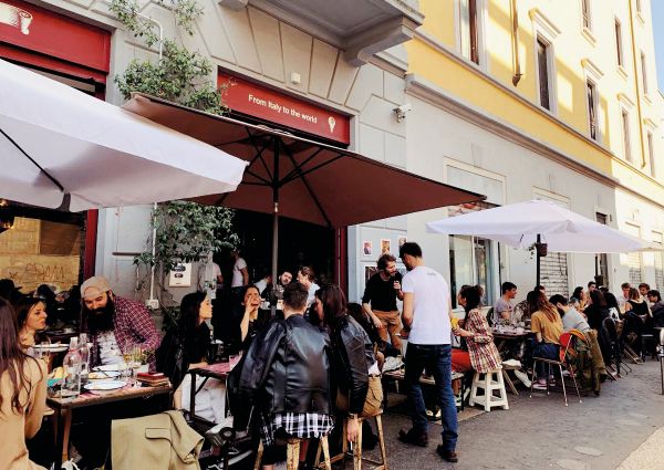 Bar affollato all'aperto. Clienti seduti, ombrelloni aperti. Flavio Angiolillo, barman italiano.