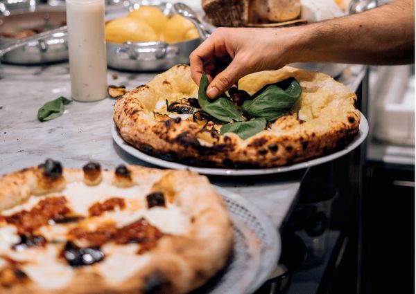 Pizza gourmet Cocciuto. Mani che aggiungono basilico fresco. Cucina fusion, eleganza e sapori unici.