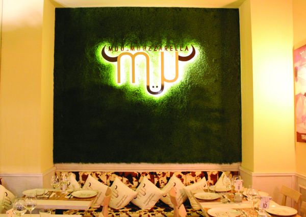 Interni di Muu Muuzzarella: logo luminoso su parete verde, tavola imbandita. Eccellenza locale e format innovativo.