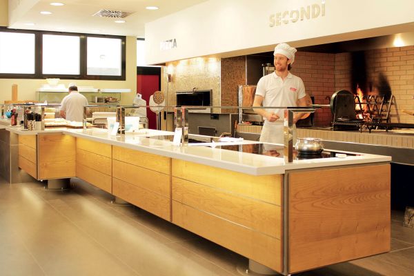 Ristorazione Camst: cuoco sorridente in mensa aziendale. Forno pizza e banco self-service.