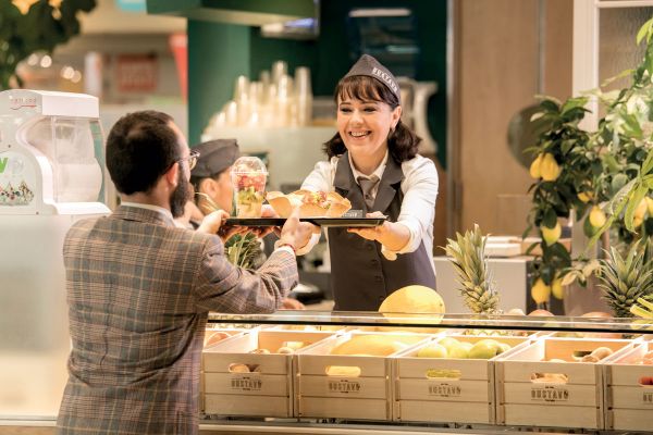 Camst: Ristorazione sana e deliziosa. Donna sorridente serve cibo e frutta fresca al cliente.