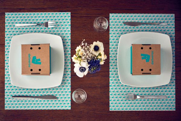 Tavola imbandita con consegna Deliveroo. Piatti, posate, fiori e box Deliveroo.