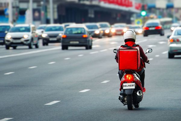 Consegna cibo: rider in moto con box rosso nel traffico cittadino. Delivery dopo le 18:00.