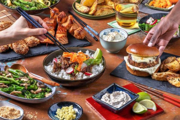 Tavola imbandita con piatti HelloFresh: burger, riso, verdure, spiedini. Nuovi consumi alimentari italiani.