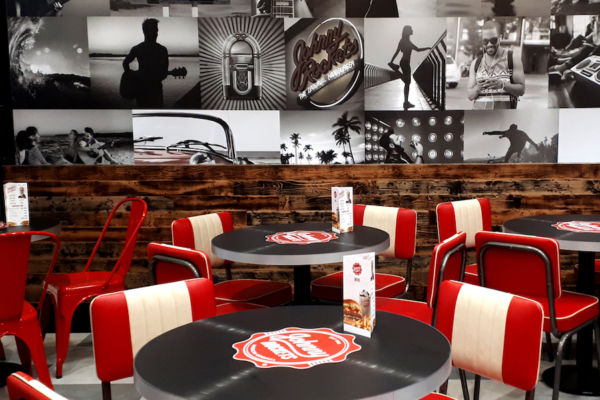 Interni Johnny Rockets Curno. Tavoli neri con logo, sedie rosse e decorazioni murali vintage. Ristorante americano anni '50.
