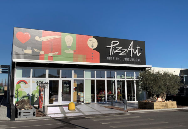 Esterno PizzAut, ristorante inclusivo. Design moderno, vetrate ampie, insegna Nutriamo l'inclusione. Qualità dell'aria e inclusione sociale.