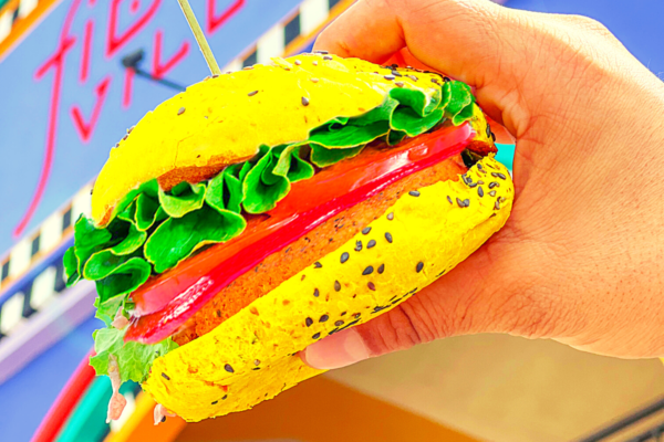 Flower Burger: mano che tiene un colorato hamburger vegano con pane giallo e verdure fresche.