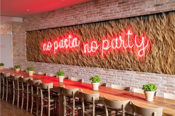 Interno ristorante miscusi: tavolo lungo, scritta neon no pasta no party, parete mattoni.