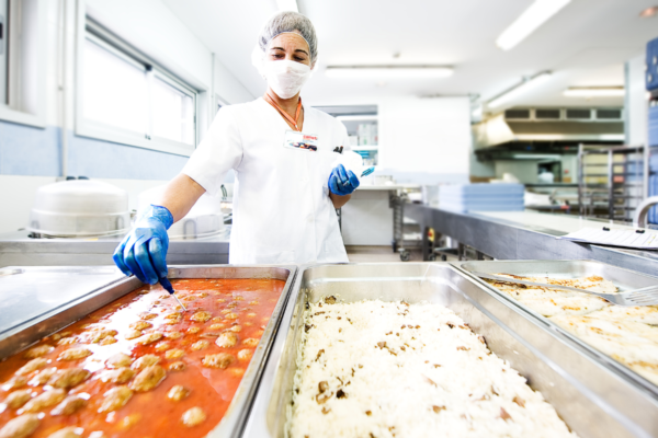 Ristorazione collettiva: addetta mensa con mascherina serve il pranzo. Cucina professionale.