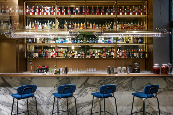 Interno bar Obicà: scaffali con bottiglie, bancone in marmo e sgabelli blu. Design elegante.